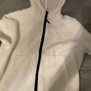 Vit hoodie från C.P. Company - Säljer en snygg vit hoodie från C.P. Company i storlek M. Den har en dragkedja framtill och en cool detalj med glasögon i huvan. Perfekt för höst och vinter, och den är i nyskick. Superbekväm och stilren, passar till alla tillfällen!