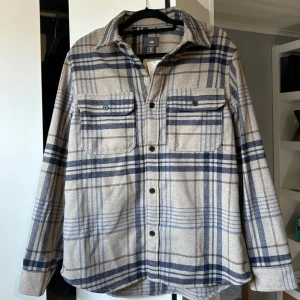 Rutig overshirt från H&M - Säljer en snygg rutig overshirt från H&M i storlek M. Den är beige med blå och grå rutor, har knappar framtill och två bröstfickor med knappar. Perfekt för höst och vår, och kan bäras som en lätt jacka eller som en skjorta. Materialet känns mjukt och bekvämt. Aldrig använd, lapp sitter kvar. Pris går att diskuteras
