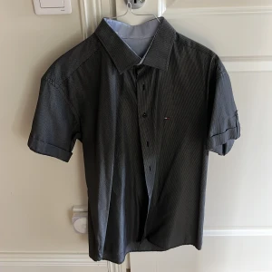 Svart och vit rutig skjorta från Tommy Hilfiger - Säljer en snygg svart och vit rutig skjorta från Tommy Hilfiger i storlek L. Skjortan är kortärmad och har knappar framtill. Perfekt för både vardag och fest! Den är i bra skick och har en klassisk design med en liten logga på bröstet. Materialet känns bekvämt och är av hög kvalitet.