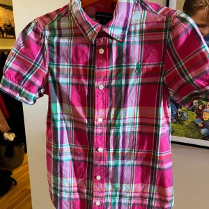Rutig skjorta från Ralph Lauren - Säljer en superfin rutig skjorta från Ralph Lauren i storlek 8. passar tjej i ålder 7-8. Skjortan har en färgglad design med rutor i rosa, grönt, vitt och rött. Den har korta ärmar och knappar hela vägen ner. Perfekt för både vardag och fest!