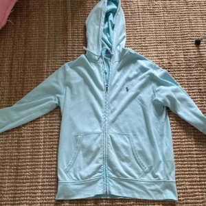 Ljusblå hoodie från Ralph Lauren - Säljer en superfin ljusblå hoodie från Ralph Lauren. Den har en dragkedja framtill och två fickor. Perfekt för vår och sommar! Den är i bra skick och väldigt bekväm. Passar både till vardags och träning.
