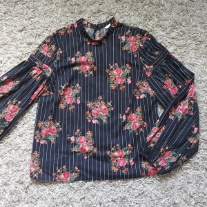 Blommig långärmad topp med randigt mönster - Säljer en superfin långärmad topp/blus  i storlek 36. Den är svart med vita smala ränder och har ett vackert blommigt mönster i rosa och grönt. Toppen har en hög hals med en liten knapp i nacken och volangdetaljer på ärmarna. Perfekt för både vardag, kontor och fest.
