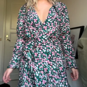 Blommig omlottklänning - Säljer en superfin blommig omlottklänning med lång ärm. Klänningen har en v-ringad hals och ett vackert mönster med rosa och gröna blommor på en svart bakgrund. Perfekt för vår och sommar! 💐Den saknar en knapp❤️ annars är den knappt använd