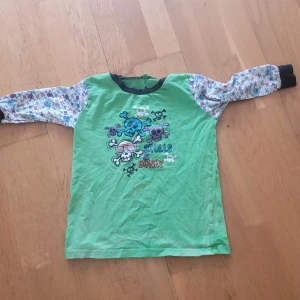 Grön långärmad tröja med dödskallemotiv - Säljer en cool grön långärmad tröja med dödskallemotiv och texten 'Skulls & Bones' på framsidan. Ärmarna är mönstrade med blommor och har svarta muddar vid ärmsluten. Perfekt för vardagsbruk eller lek!