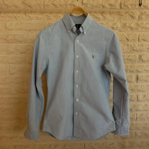 Ljusblå skjorta från Ralph Lauren - Säljer en snygg ljusblå skjorta från Ralph Lauren i storlek S. Skjortan är långärmad och har en klassisk krage med knappar. Den är tillverkad i bomull och har en broderad logga på bröstet. Perfekt för både vardag och fest!