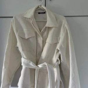 Vit manchesterjacka med bälte - Säljer en snygg vit manchesterjacka. Jackan har långa ärmar och ett bälte i midjan för en fin passform. Den har också två bröstfickor med lock och knappar. Perfekt för vår och höst!