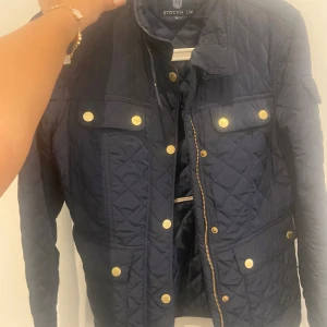 Mörkblå quiltad jacka från Stockh LM - Säljer en snygg mörkblå quiltad jacka från Stockh LM. Jackan har en dragkedja framtill och flera guldknappar som detaljer. Den har långa ärmar och fyra fickor med knappar på framsidan. Perfekt för höst och vår!