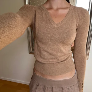Zara topp - En långärmad v-ringad topp från Zara i en camel-brun/beige färg. Toppen har knappar i ärmarna och är i en slim-fit. Använd men fortfarande väldigt snygg!  Är lite nopprig på sina ställen men går att ta bort med en noppborttagare.