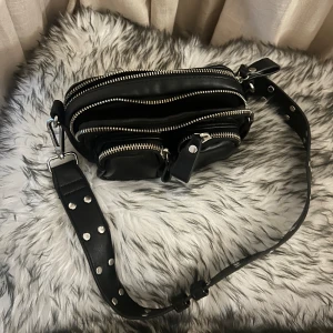 Svart axelväska från Gina Tricot - Säljer en snygg svart axelväska från Gina Tricot. Väskan har flera dragkedjeförsedda fack och en justerbar axelrem med nitar. Perfekt för både vardag och fest!