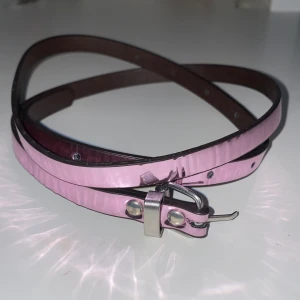 Rosa smalt bälte - Säljer ett smalt rosa bälte i skinn med en silverfärgad spänne. Bältet är i nyskick och har en glansig yta som ger en snygg touch till vilken outfit som helst. Perfekt för att piffa upp dina jeans eller klänningar!