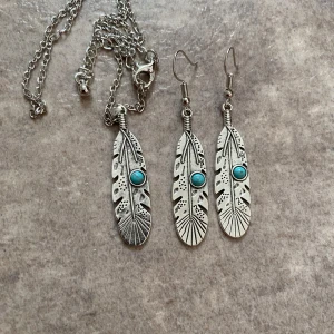 Boho smycken  - Ett set med tillhörande örhängen och halsband i rostfritt stål format som fjäder med turkos sten. Halsbandet är 40 cm långt och går att justera. Örhängen är 5,5 cm långa. Helt nya.