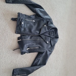 Svart skinnjacka med dragkedja och bältesdetaljer - Säljer en snygg svart skinnjacka i storlek 158. Jackan har en cool bikerstil med dragkedja framtill och två fickor med dragkedjor. Den har också bältesdetaljer vid midjan. Perfekt för höst och vår! 🖤