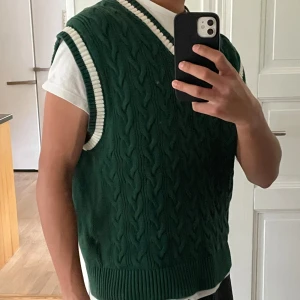 Grön stickad väst - Säljer en snygg grön stickad väst från H&M i storlek S. Den har ett klassiskt kabelstickat mönster och vita ränder vid ärmhålen och halsringningen. Perfekt för lager-på-lager under höst och vår. Västen är endast använd ett fåtal gånger.