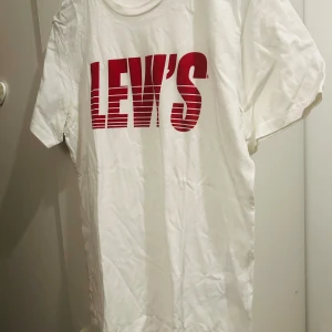 Vit t-shirt från Levi's - Säljer en klassisk vit t-shirt från Levi's med deras ikoniska röda logga tryckt på framsidan. T-shirten är i bomull och har en normal passform med korta ärmar. Perfekt för en avslappnad stil!