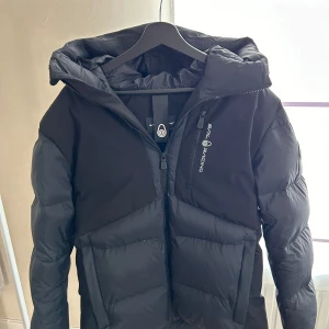 Svart dunjacka från Sail Racing - Säljer en snygg och varm svart dunjacka från Sail Racing. Jackan har en dragkedja framtill och en praktisk huva. Perfekt för kalla vinterdagar. Den är i nyskick och redo för nya äventyr!