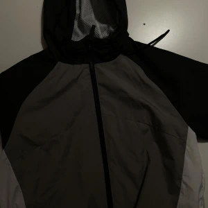 Grå och svart vindjacka från Primark - Säljer en grå och svart vindjacka från Primark. Jackan har en dragkedja framtill och en huva med nätfoder. Perfekt för blåsiga dagar och passar bra till vardags eller lättare utomhusaktiviteter.