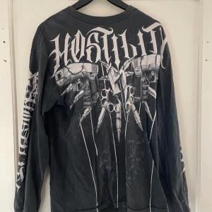 Svart långärmad tröja från Hostility - Säljer en cool svart långärmad tröja från Hostility med ett unikt tryck i vitt. Tröjan har ett edgy motiv med detaljer som ger en tuff look. Perfekt för dig som vill sticka ut!