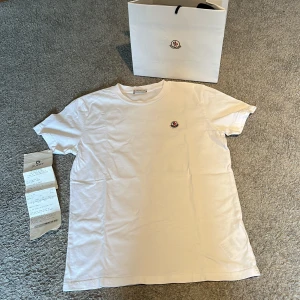Vit Moncler T-shirt - Säljer den här riktigt snygga vita t-shirten från Monclear som jag vuxit ur. Tshirten är i storlek 14A vilket är storlek 164 som förklarat på dem två sista bilderna😁Bara använt tröjan några gånger så den är i helt nytt skick👍Påse och kvitto ingår såklart💯Nypris är 1800kr✅✅✅