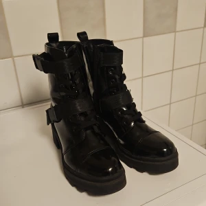Svarta lackade boots med snörning och spännen - Säljer ett par svarta, glansiga boots i lack med både snörning spännen. De har en rejäl sula som ger bra grepp och en edgy look. Perfekta för höst och vinter när du vill ha något som både är snyggt och praktiskt. Passar till både jeans och k