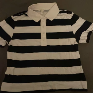 Randig pikétröja i svart och vit - Säljer en klassisk randig pikétröja i Blå och vit. Den har en krage och knappar framtill, perfekt för en avslappnad men stilren look. Tröjan är i storlek S och passar både till vardags och lite mer uppklädda tillfällen.