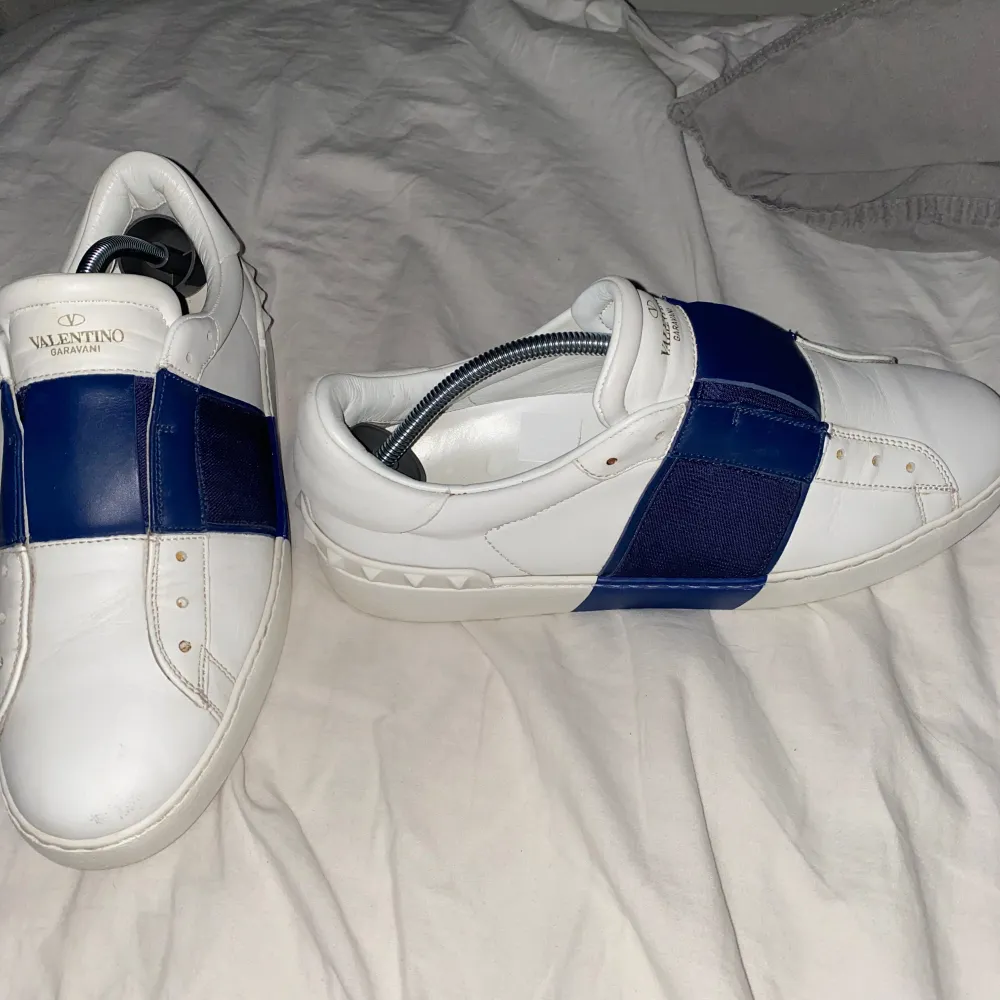 Säljer ett par snygga Valentino sneakers i vitt med en cool blå rem över ovansidan. Skorna är i skinn och har en stilren design med diskreta nitar på sidorna. Perfekta för både vardag och fest!. Skor.