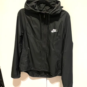 Svart vindjacka från Nike - Säljer en snygg och praktisk svart vindjacka från Nike. Jackan har en dragkedja framtill och en justerbar huva. Perfekt för blåsiga dagar och passar till både träning och vardag. Den är i nyskick, passar XS och S