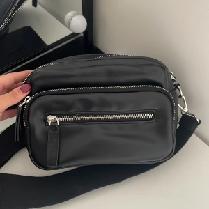 Svart axelväska med dragkedjor - Säljer svart axelväska i syntetmaterial. Väskan har flera praktiska fack med dragkedjor och en justerbar axelrem. Perfekt för vardagsbruk eller när du är på språng. Stilren och enkel att matcha med olika outfits!