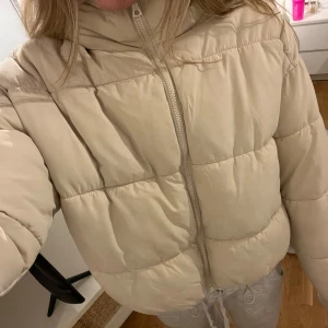 Beige pufferjacka från Monki - Säljer en supermysig beige pufferjacka från Monki i storlek M som är croppad. Jackan har en dragkedja framtill och en skön huva. Perfekt för kyliga höst- och vinterdagar. Den är i mycket bra skick.