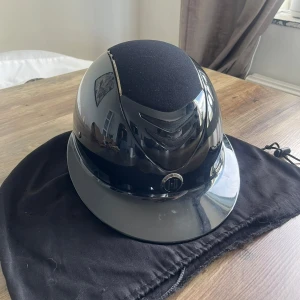 Svart ridhjälm - Snygg och stilren svart ridhjälm med glansig yta och matt tygdetalj på toppen. Perfekt för ridning och ger ett elegant intryck. Hjälmen ser ut att vara i mycket bra skick och kommer med en praktisk förvaringspåse.