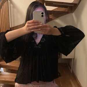 Blus - Säljer en elegant svart blus med vackra pärldetaljer vid halsringningen. Blusen har långa, vida ärmar och ett lätt transparent material som ger en luftig känsla. Perfekt för både fest och vardag! Defekt är att öglans där man sätter fast knappen har lossnat men inget man behöver knäppa och enkelt att fixa (bild 3)