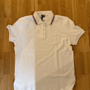  pikétröja Moncler - Säljer en snygg vit pikétröja från Moncler i nytt skick. Den har en klassisk krage med röd och svart randig detalj och Moncler-logga på ärmen.