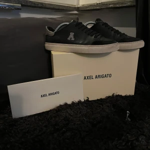 Svarta sneakers från Axel Arigato - Säljer ett par sjukt stilrena pjucks från Axel Arigato i läder med bokstaven A på sidan som en unik detalj. Perfekta för både vardag och fest! Även väldigt bra för kommande årstider då de även tål kyla. Skorna är i andvänt skick och allt og finns kvar, dvs kvitto, box och medföljande saker. 
