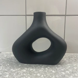 Svart dekorativ vas från H&M Home - Snygg och modern svart vas från H&M Home. Den har en unik, skulptural form med ett stort hål i mitten, vilket gör den till ett perfekt dekorativt inslag i hemmet. Passar utmärkt på ett bord eller en hylla för att ge rummet en stilren touch.
