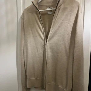 Beige tröja med dragkedja från These Glory Days - Säljer en stilren beige tröja från These Glory Days i storlek M. Tröjan har en dragkedja framtill och är perfekt för både vardag och lite finare tillfällen. Den är i mycket bra skick!