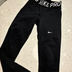 Svarta Nike Pro leggings - Säljer ett par svarta Nike Pro leggings i storlek M. De är tillverkade i Dri-FIT-material som håller dig torr och bekväm under träning. Leggingsen har en elastisk midja med Nike-logga och en liten vit Swoosh på benet. Perfekta för gymmet eller löprundan!