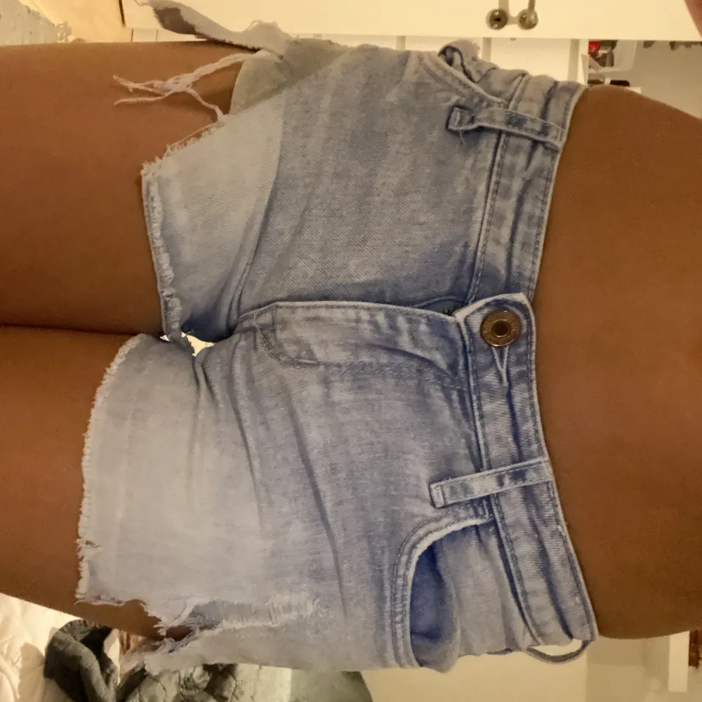 Säljer ett par ljusa jeansshorts i slitet skick med råa kanter och slitningar. Perfekta för sommardagar och festivaler! De har en knapp och dragkedja framtill samt klassiska fem fickor. Passar perfekt till en avslappnad stil.. Shorts.