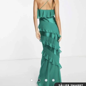 Grön långklänning med volanger - Fantastisk grön balklänning med volanger. Den har en elegant oneshoulder-design. Klänningen är i ett mjukt och följande material som ger en vacker siluett. Använd 1 gång på bal, därav små fläckar nertill