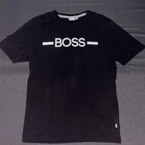 Svart t-shirt från Hugo Boss - Säljer en stilren svart t-shirt från Hugo Boss i storlek M. Den har en vit BOSS-logga på bröstet som ger en snygg kontrast. Perfekt för både vardag och fest. T-shirten är i bra skick och gjord av mjukt material för en bekväm passform.