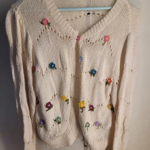 Beige stickad kofta med blommor - Superfin beige stickad kofta med färgglada blommor broderade över hela. Den har en v-ringad design och knäpps med söta knappar i olika färger. Perfekt för vår och sommar när du vill ha något lätt och luftigt men ändå mysigt. Passar både till jeans och klänning!