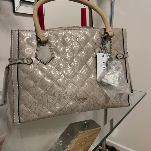Beige handväska från Guess - Säljer en snygg beige handväska från Guess i nytt skick. Väskan har ett quiltat mönster med Guess-loggan och är gjord i syntetmaterial. Den har två handtag och en dragkedja upptill. Perfekt för både vardag och fest!