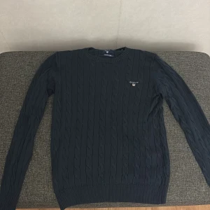 Svart kabelstickad tröja från GANT - Säljer en snygg svart kabelstickad tröja från GANT i premium bomull. Tröjan har en klassisk design med ribbade muddar och rund hals. Perfekt för både vardag och lite finare tillfällen. Passar bra till höst och vinter!