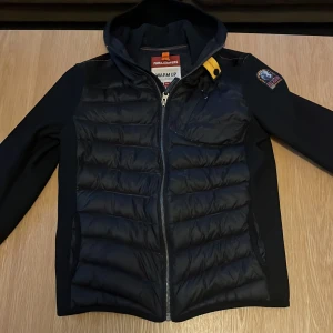 Svart jacka från Parajumpers - Säljer en snygg svart jacka från Parajumpers. Den har en quiltad framsida och baksida med dragkedja och en cool patch på ärmen. Perfekt för höst och vår med sin stilrena design och funktionella detaljer. Jackan har en huva och är i mycket bra skick. Passar perfekt för både vardag och äventyr!