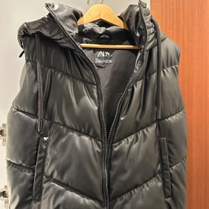 Svart dunväst från Zara - Säljer en snygg svart dunväst från Zara. Den har en dragkedja framtill och en mysig huva med dragsko. Perfekt för höst och vinter när du vill hålla dig varm och stilren. Västen är i mycket bra skick och passar perfekt för både vardag och fest.