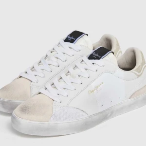 Vita sneakers från Pepe Jeans - Snygga vita sneakers från Pepe Jeans i skinn med detaljer i beige och guld. De har en klassisk snörning och en bekväm passform. Perfekta för både vardag och festliga tillfällen. Sulan är lätt smutsad för en cool, använd look.