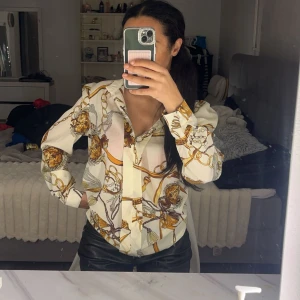 Mönstrad blus med kedjedetaljer - Säljer en snygg blus med ett unikt kedjemönster i guld och brunt på en vit bakgrund. Blusen har långa ärmar och en klassisk krage. Perfekt för både vardag och fest! 🖤