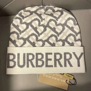 Vit och grå mössa från Burberry - Snygg och stilren mössa från Burberry i vitt och grått med deras ikoniska logga. Den är stickad och perfekt för att hålla dig varm under kyliga dagar. Passar både till vardags och lite finare tillfällen. Ett måste för modeälskare!