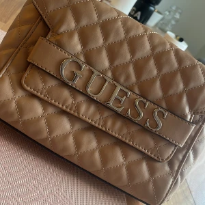 Beige quiltad axelväska från Guess - Säljer en snygg beige axelväska från Guess i mycket bra skick. Väskan är quiltad och har en stilren design med silverdetaljer och kedjehandtag. Perfekt för både vardag och fest! 👜✨