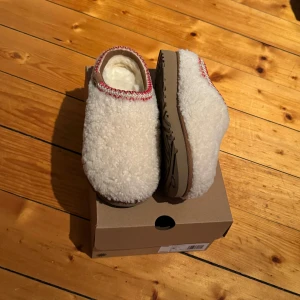 UGG tasman maxi curly - Mysiga tofflor från UGG i ett fluffigt material som ser ut som päls. De har en snygg röd detalj runt kanten och en stabil sula. Perfekta för att hålla fötterna varma under kalla dagar hemma. Skorna är helt nya och aldrig använda. Finns i storlekarna 37, 38 och 39.