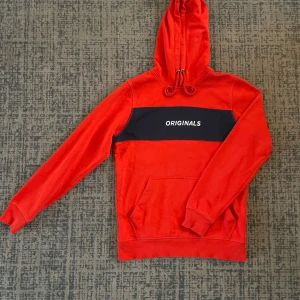 Röd hoodie från Jack & Jones - Säljer en snygg röd hoodie från Jack & Jones med texten 'ORIGINALS' på bröstet. Den har en klassisk design med känguruficka och justerbar huva. Perfekt för en avslappnad stil eller kyliga dagar. Passar både till vardags och chillkvällar. 🧡