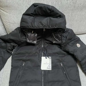 Svart dunjacka från Moncler - Säljer en snygg svart dunjacka från Moncler i mycket bra skick. Jackan har en dragkedja framtill och en huva för extra värme. Den är perfekt för kalla vinterdagar och har Monclers ikoniska logga på ärmen. Insidan har en cool serietidningsdesign. Passar perfekt för både vardag och fest!